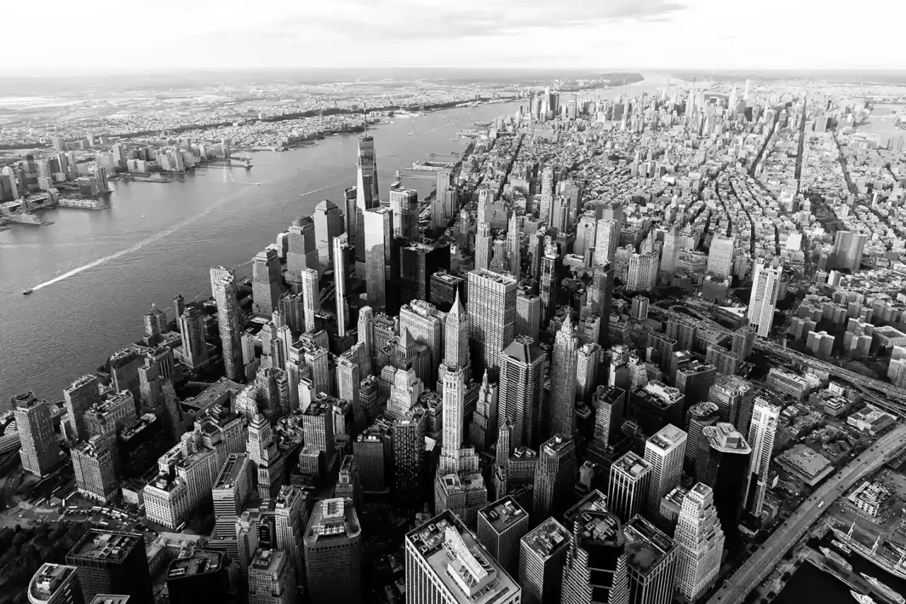 Aerial view of a densely packed urban skyline with tall skyscrapers, city streets, and a river running alongside the cityscape in black and white.