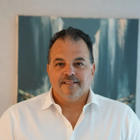 A middle-aged man with short dark hair and a goatee, wearing a white collared shirt, stands in front of a blue abstract painting.