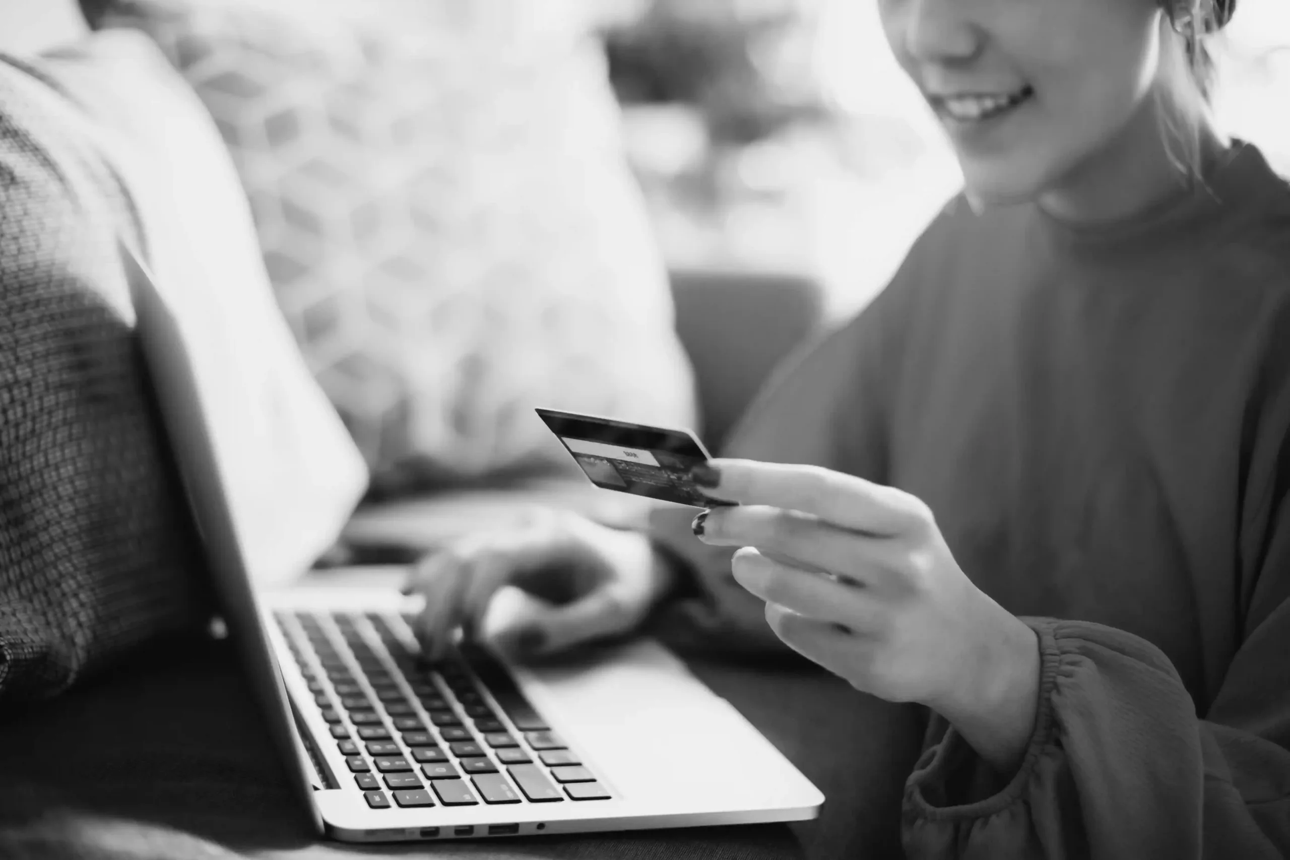 A person sits at a laptop holding a credit card in one hand, appearing to make an online purchase or payment.