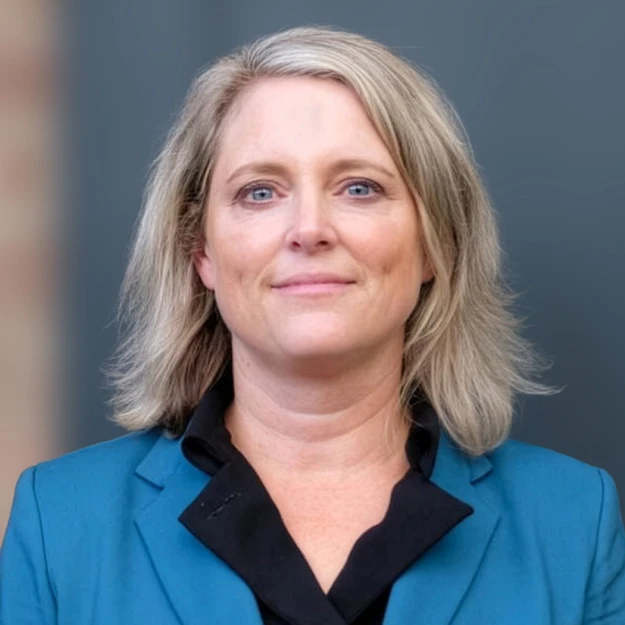 A woman with shoulder-length blonde hair is wearing a teal blazer over a black shirt, standing in front of a blurred background.