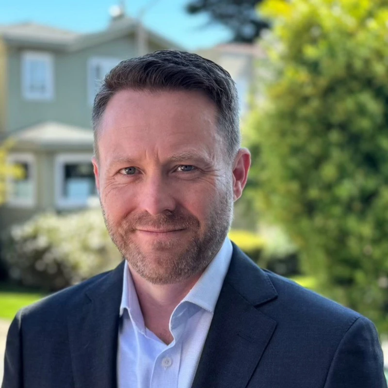 A man in a suit and collared shirt stands outdoors in front of houses and greenery, looking at the camera and smiling slightly.