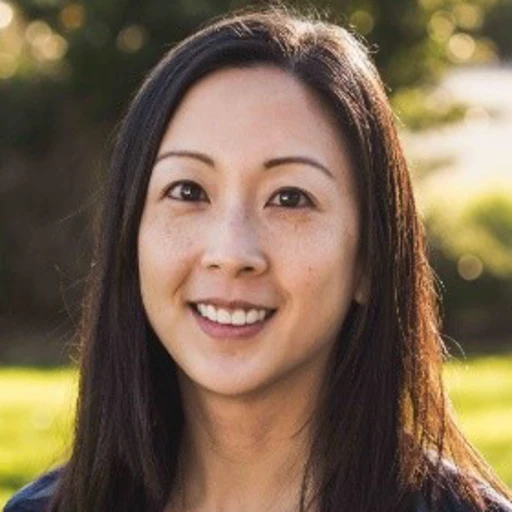 A woman with long straight hair smiles outdoors, with a blurred background of greenery and sunlight.