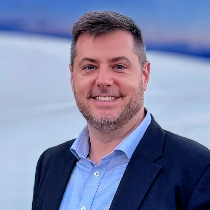 A man in a suit jacket and light blue shirt smiles at the camera while standing outside with a blurred background.
