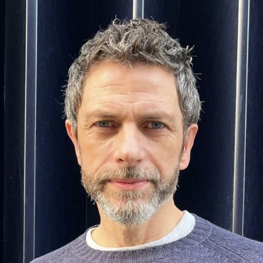A middle-aged man with short, curly gray hair and a beard, wearing a gray sweater, stands in front of a dark, vertically-striped background.