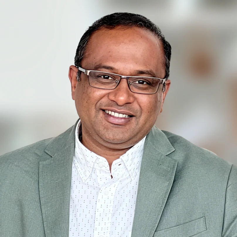 A man wearing glasses and a light gray blazer over a white patterned shirt, smiling at the camera with a blurred background.