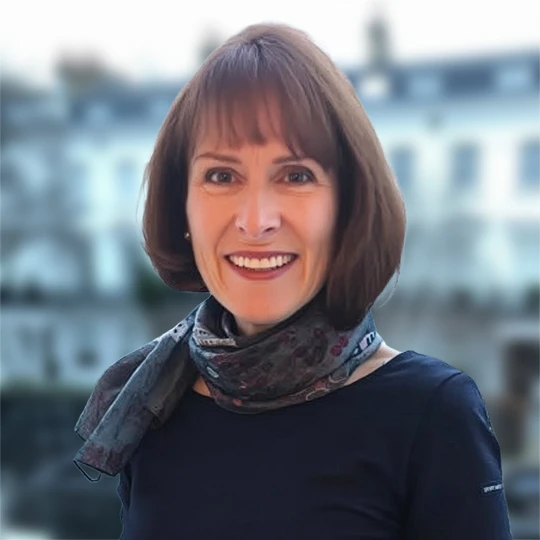 A woman with short brown hair, wearing a dark top and patterned scarf, smiles at the camera with blurred buildings in the background.