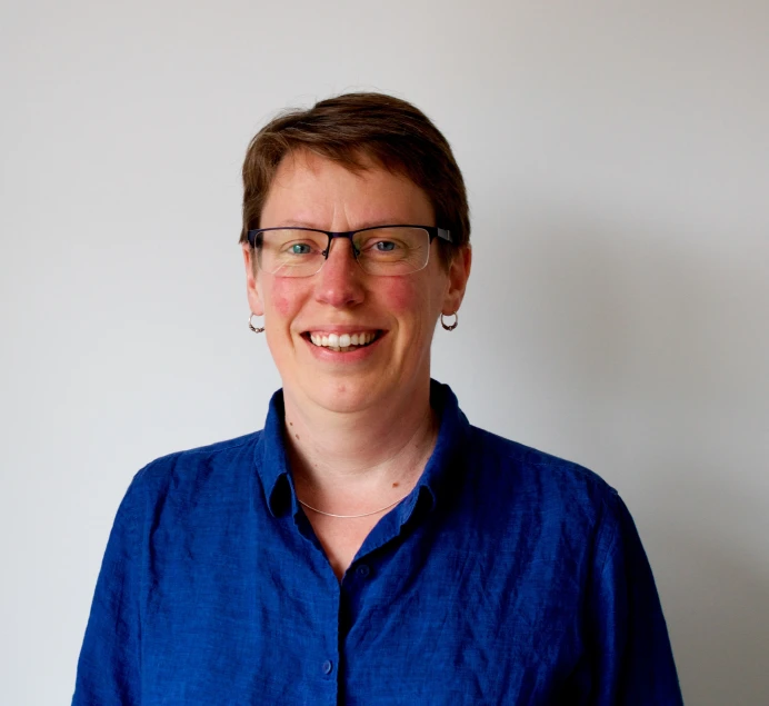 A person with short brown hair and glasses, wearing a blue collared shirt and small hoop earrings, smiling in front of a plain light background.