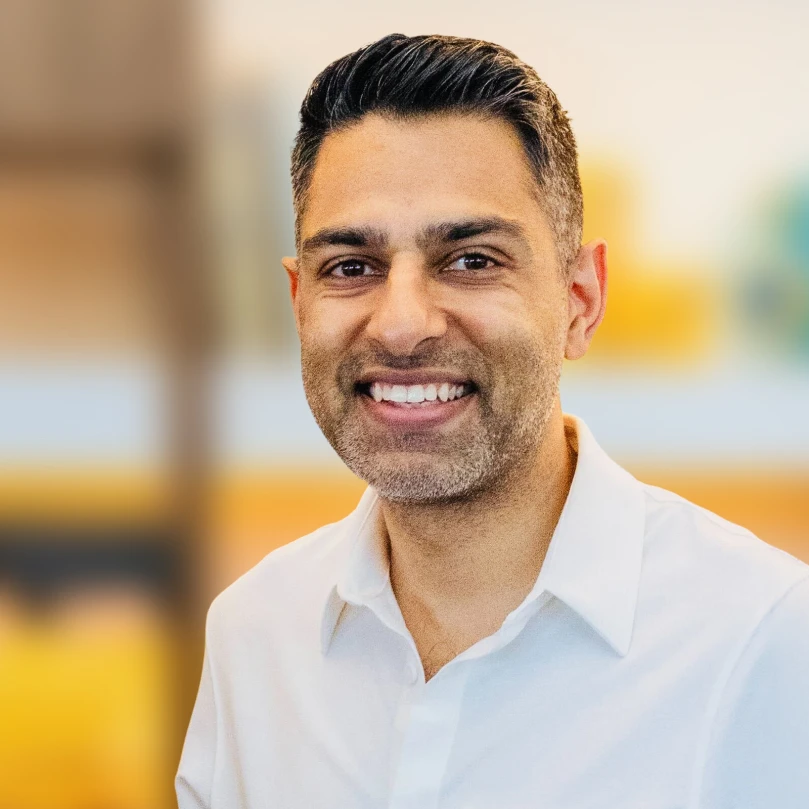 A man with short dark hair and a trimmed beard, wearing a white collared shirt, smiles at the camera against a blurred indoor background.