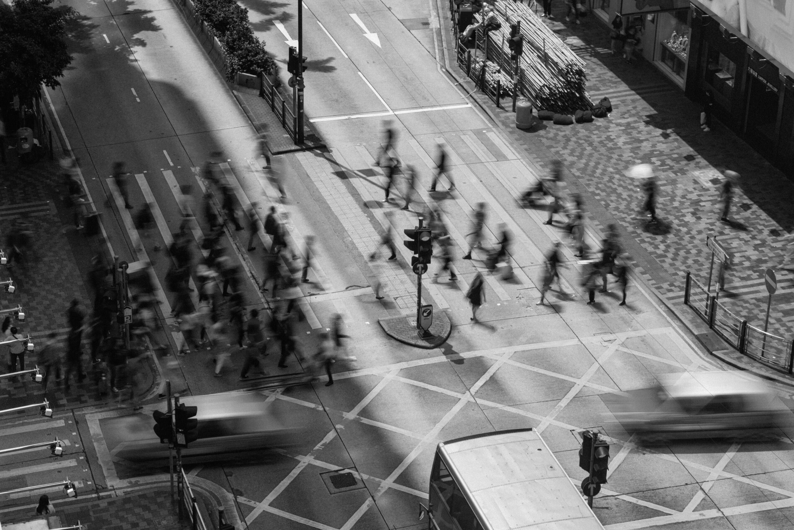 A black-and-white aerial view of a busy city intersection with blurred pedestrians crossing and vehicles moving through the street.