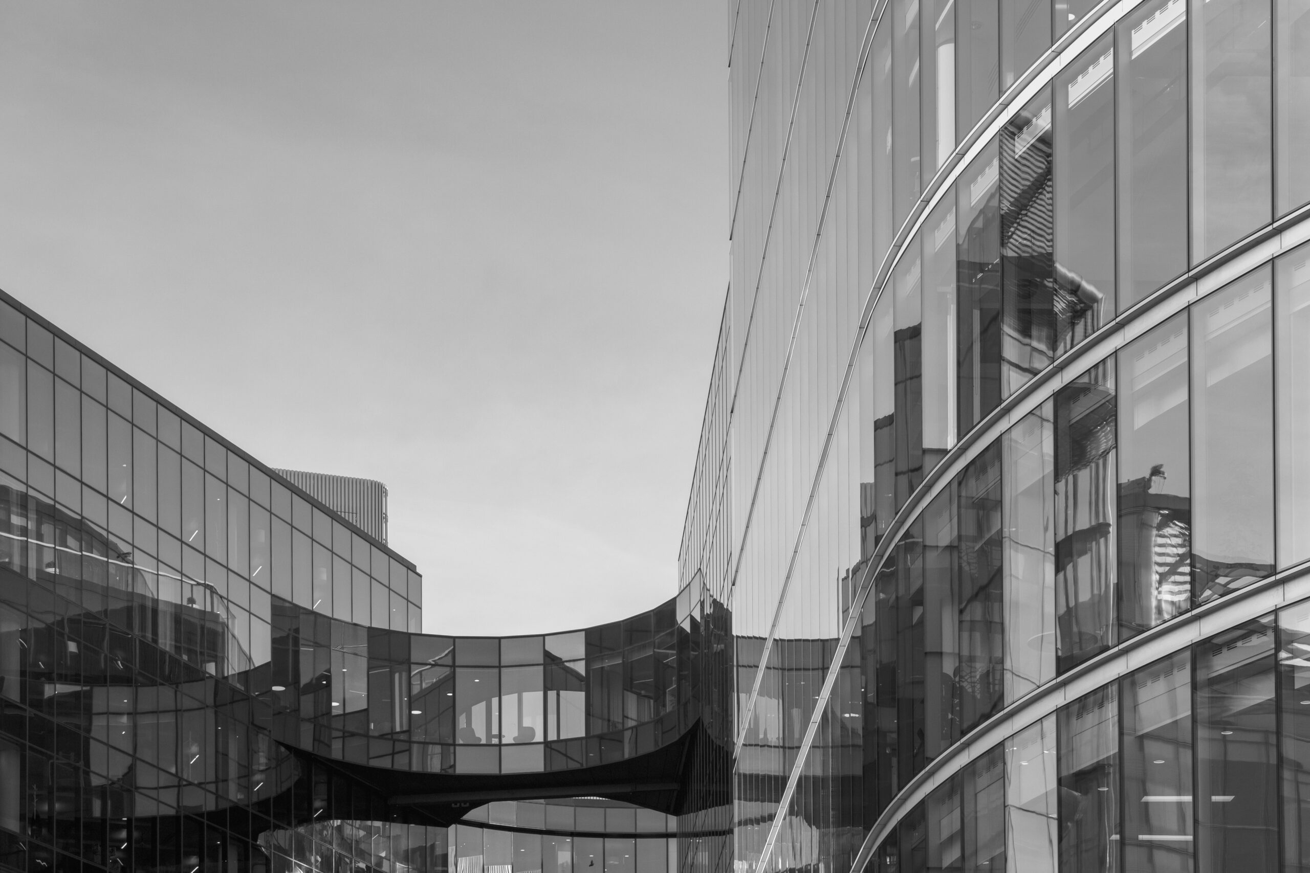 Black and white photo of modern glass office buildings connected by a skybridge; architectural reflections visible on the curved facades.