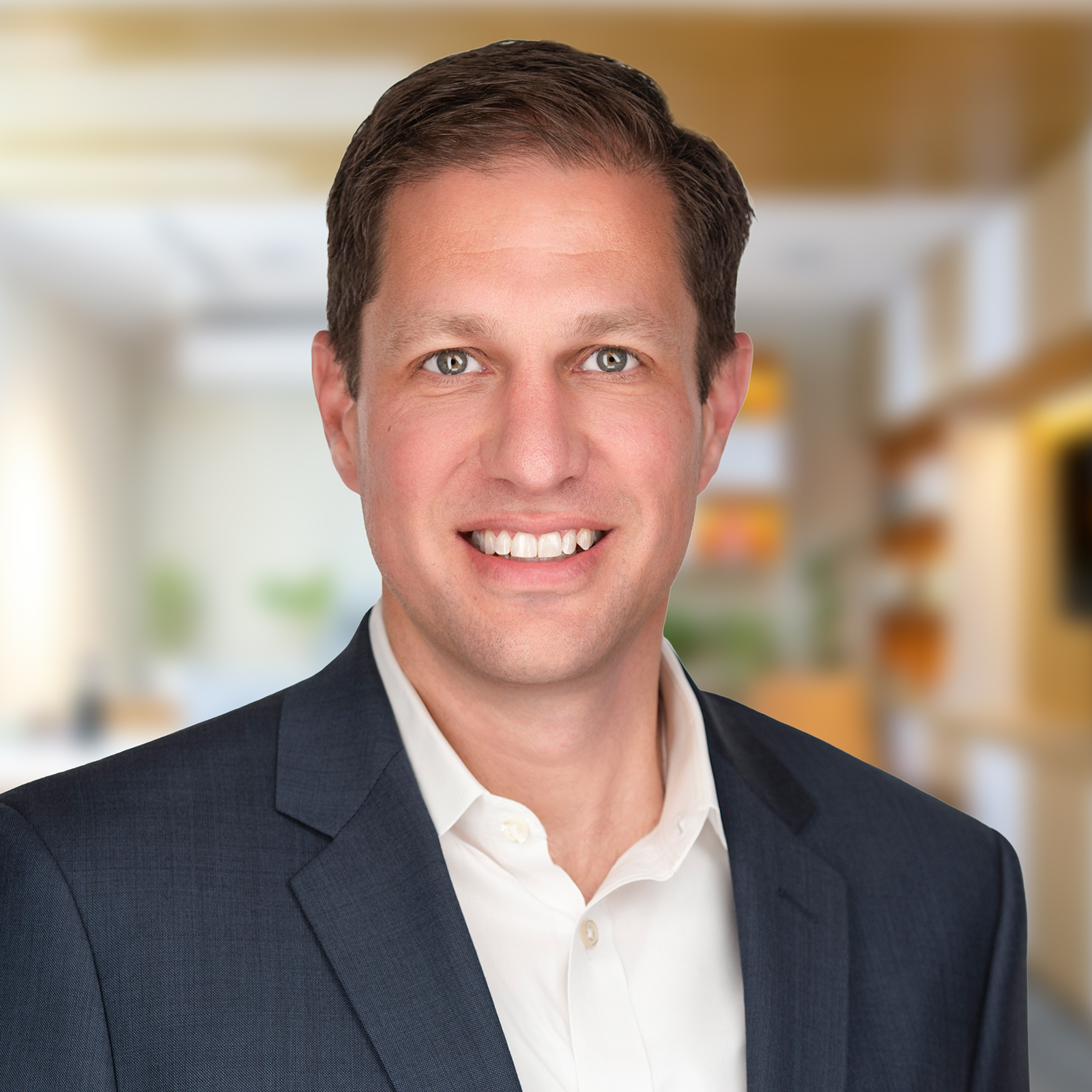 A man in a navy blue suit and white shirt is smiling, standing in a brightly lit office setting with blurred background.