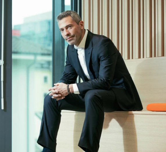 Un homme en costume noir est assis sur un banc en bois près d'une grande fenêtre et regarde la caméra en joignant les mains.