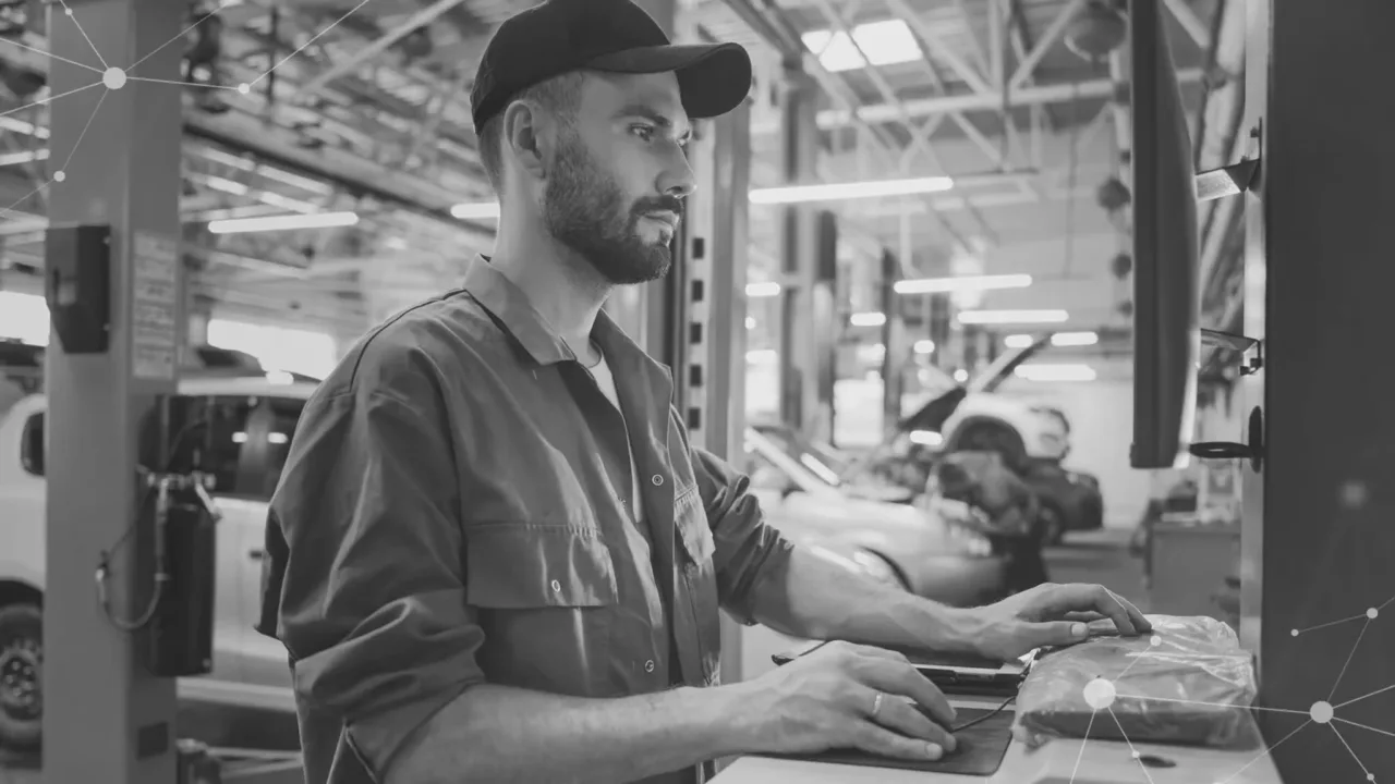 A mechanic wearing a cap and work uniform uses a computer at an automotive repair shop, with cars and equipment visible in the background.