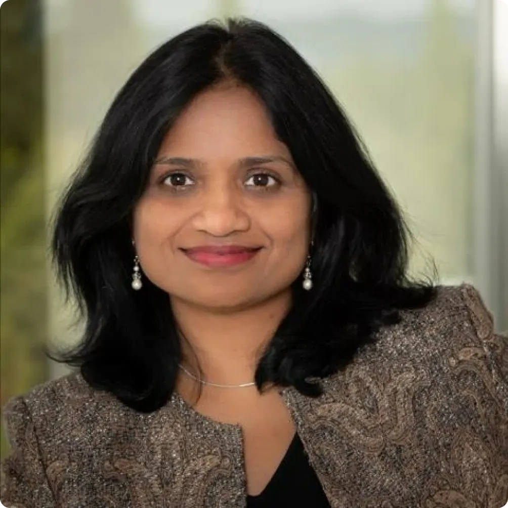 Woman with medium-length black hair, wearing a brown patterned blazer, black top, and pearl earrings, smiling at the camera with a blurred background.