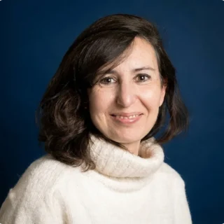 Eine Frau mit mittellangem, dunklem Haar, die einen cremefarbenen Rollkragenpullover trägt und vor einem dunkelblauen Hintergrund sanft lächelt.