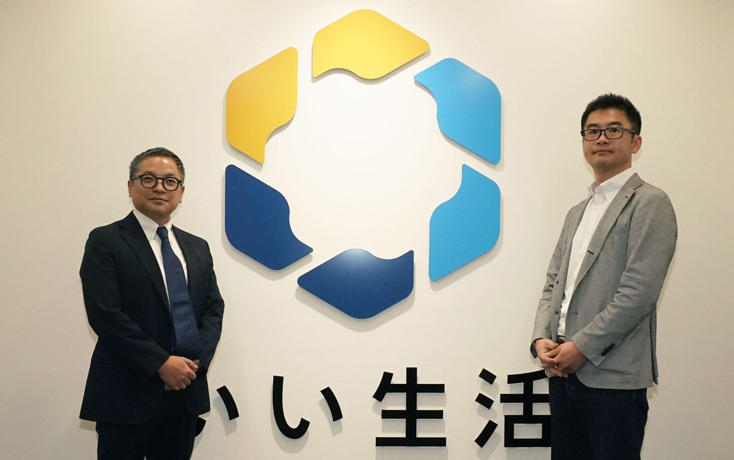 青と黄色の幾何学的なロゴとSaaSとクラウドに関連する日本語のテキストが描かれた壁の前に立つ2人の男性。.