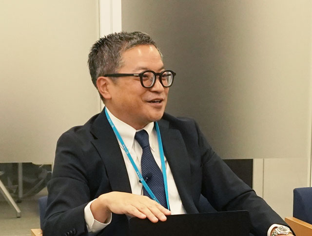 オフィスチェアに座り、笑顔で話すビジネススーツに眼鏡をかけた男性の首にはヒモが巻かれている。.