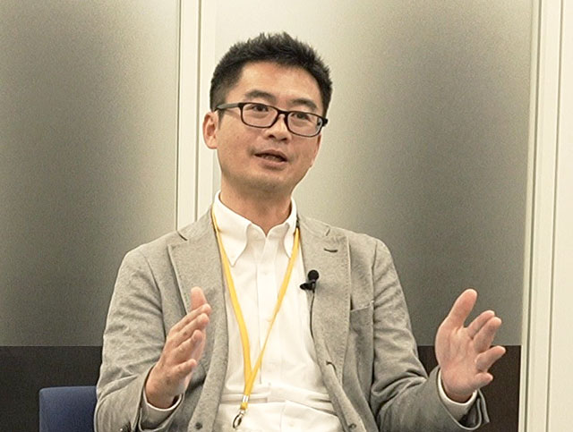 眼鏡をかけ、白いシャツを着て、薄いグレーのブレザーを着た男性が、オフィスでSaaSソリューションについて議論しているときに、両手でジェスチャーをしている。.