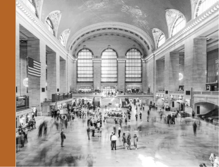 Black and white image of a busy train station interior with arched windows, high ceilings, and people walking on a large concourse. An American flag hangs on the left.