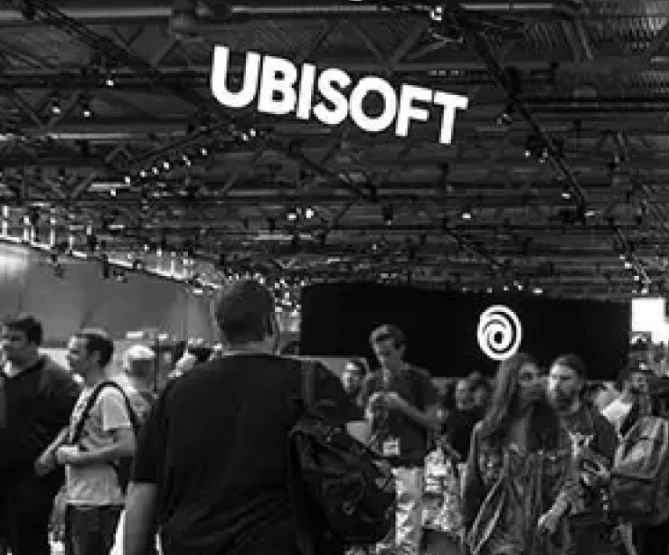 People walking near exhibits at a Ubisoft event, under a ceiling with overhead lights.